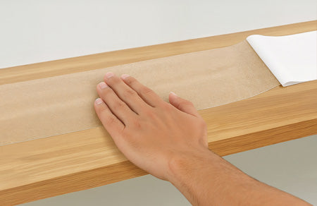 A hand sits on top of a clear stair tread being placed on a wooden step as the white backing is pulled away. 